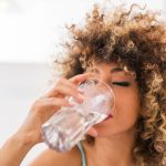 Person with curly hair drinking a glass of water indoors.
