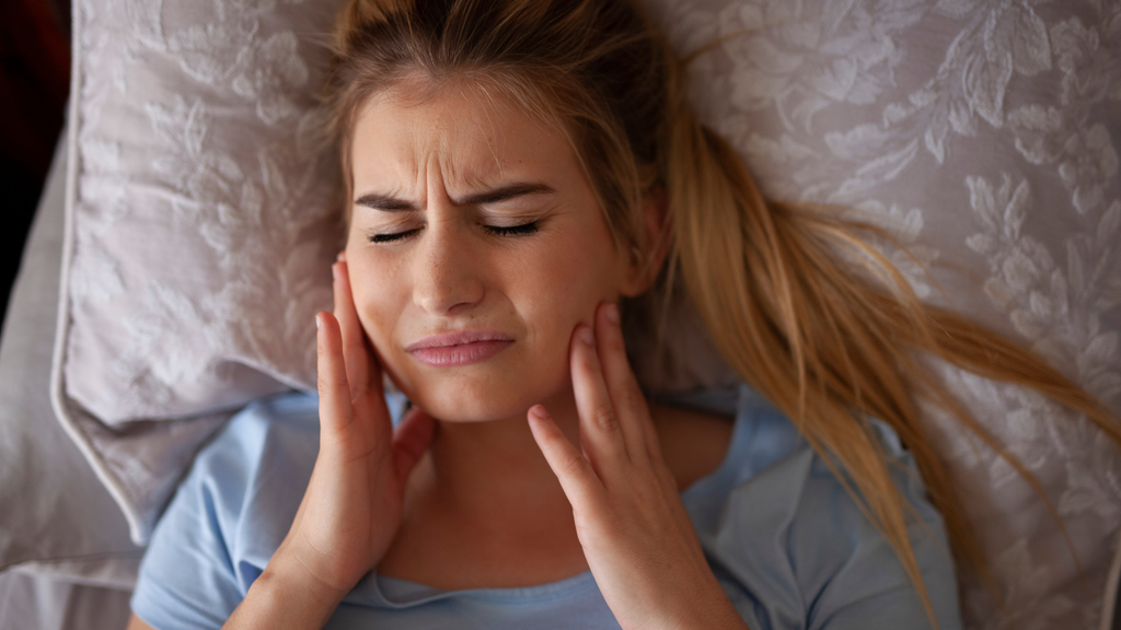 Woman lying in bed with eyes closed, touching both sides of her face and appearing to be in pain or discomfort.