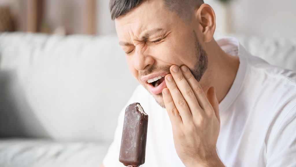 A man in a white t-shirt holds his jaw in pain while eating a chocolate ice cream bar, indicating tooth sensitivity or discomfort.